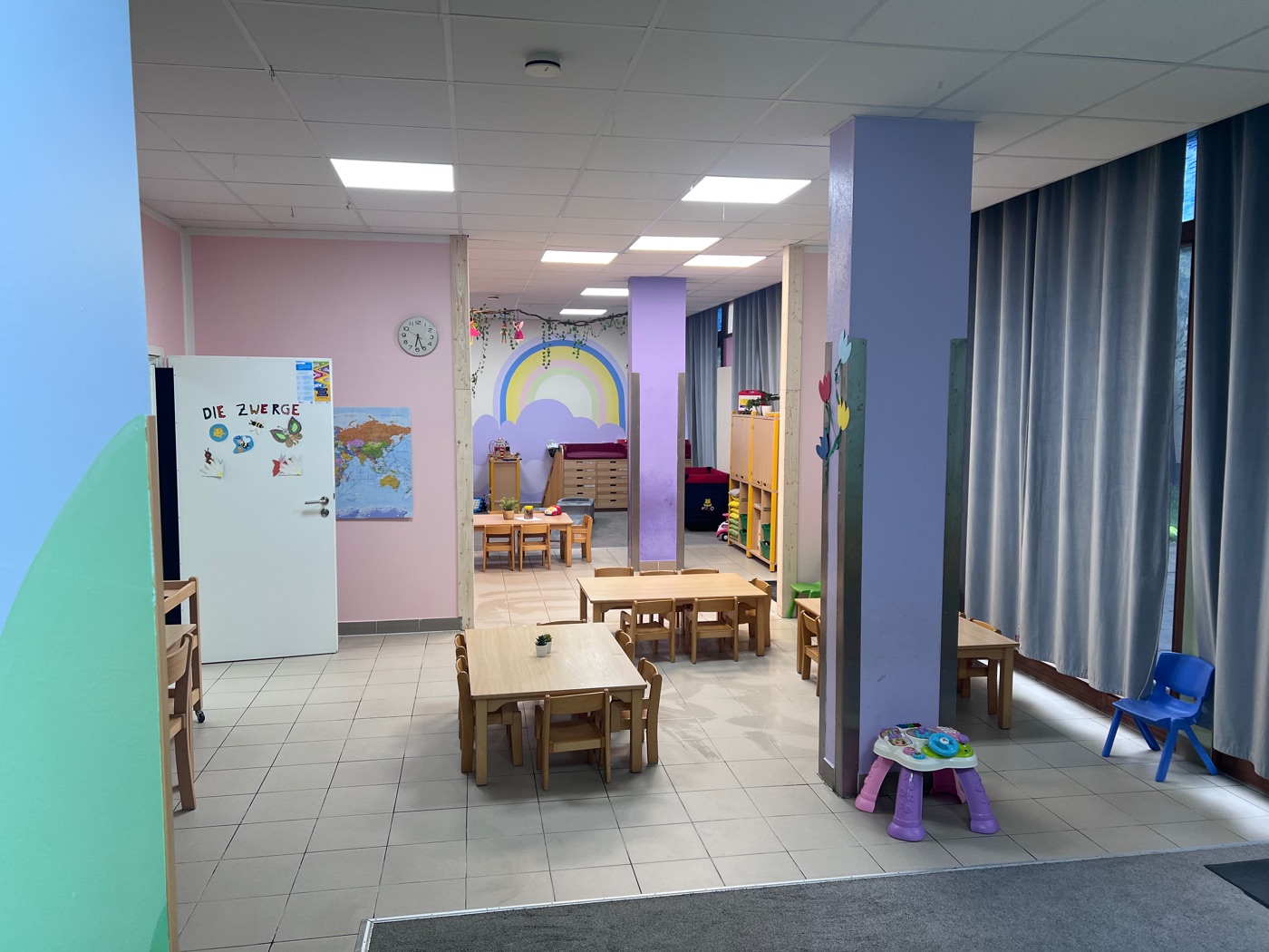 Verbundene Gruppenräume mit Kindertischen, niedrigen Regalen und Regenbogen-Wandbild
